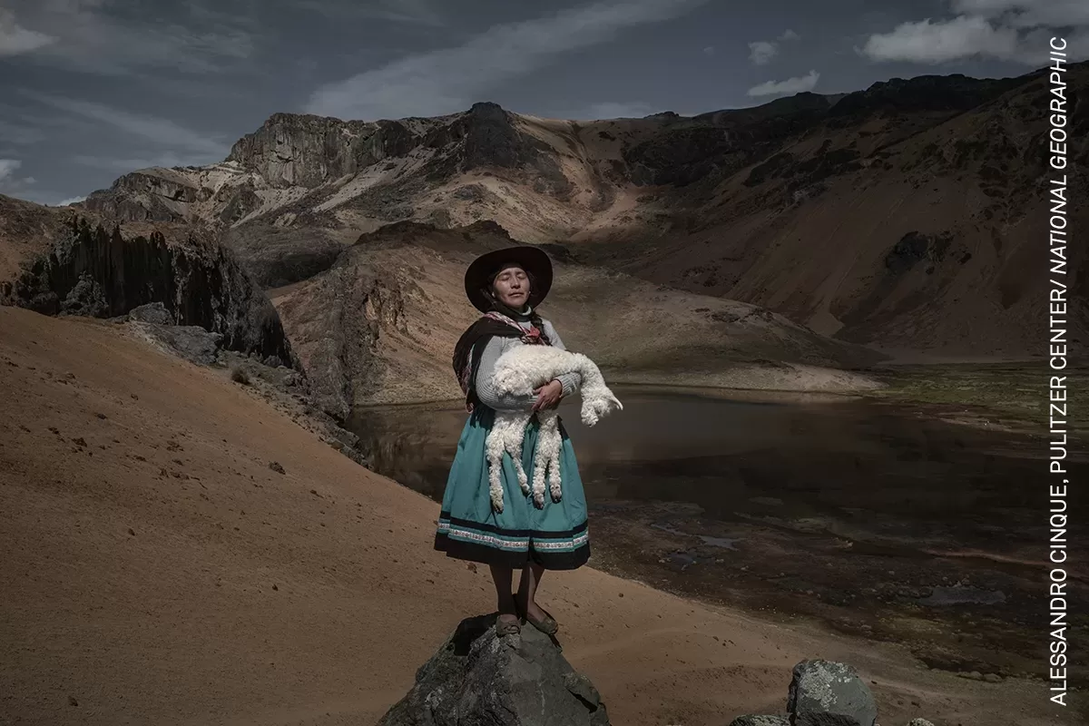 Afbeelding #GivingPhoto: Een vrouw in traditionele kledij uit Zuid-Amerika staat op een rots met een alpaca in haar armen