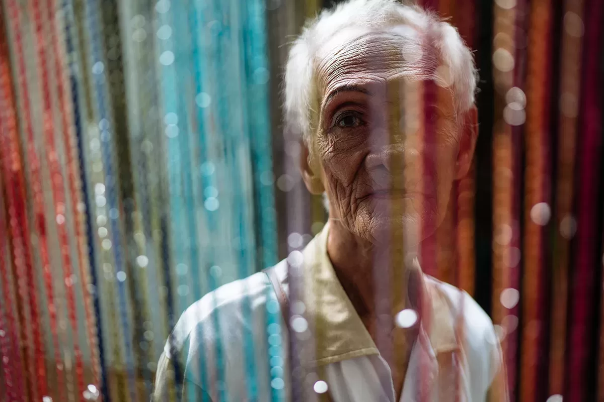 Afbeelding #GivingPhoto: een ouderen Aziatische man kijkt recht in de camera door een kleurrijk deurgordijn.