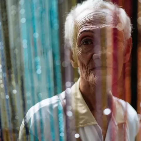 Afbeelding #GivingPhoto: een ouderen Aziatische man kijkt recht in de camera door een kleurrijk deurgordijn.